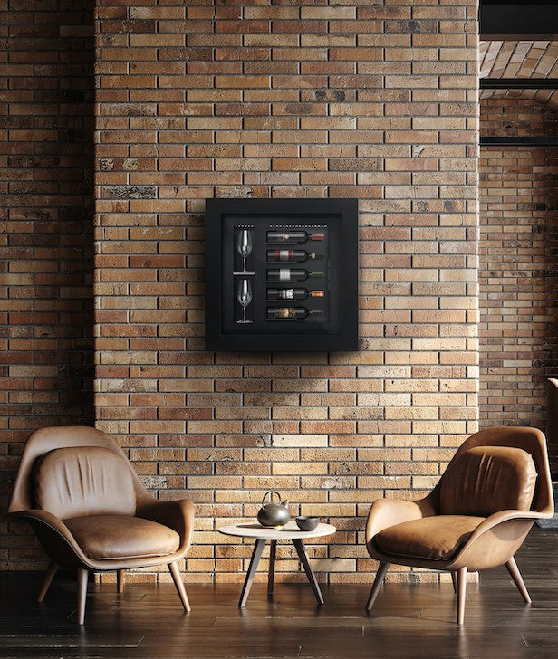A small, square black wine cabinet  containing 2 wine glasses beside 5 horizontal wine bottles mounted on a brick wall with 2 brown leather armchairs and a small round coffee table in front