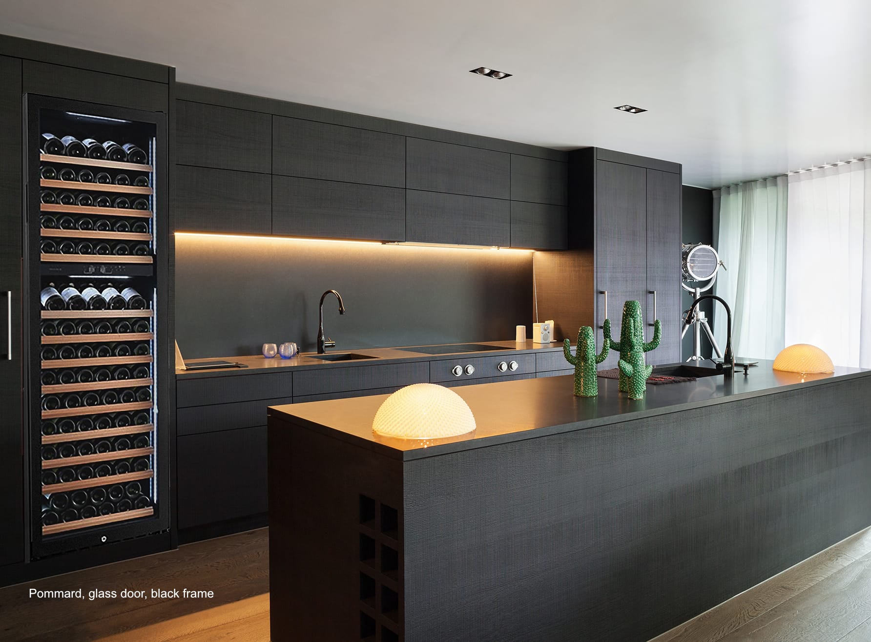 Vin Garde Pommard wine cooler in luxury kitchen setting. Glass door open showing interior with extended sliding shelves. Dark frame unit integrated between black cabinetry. Modern kitchen with light w